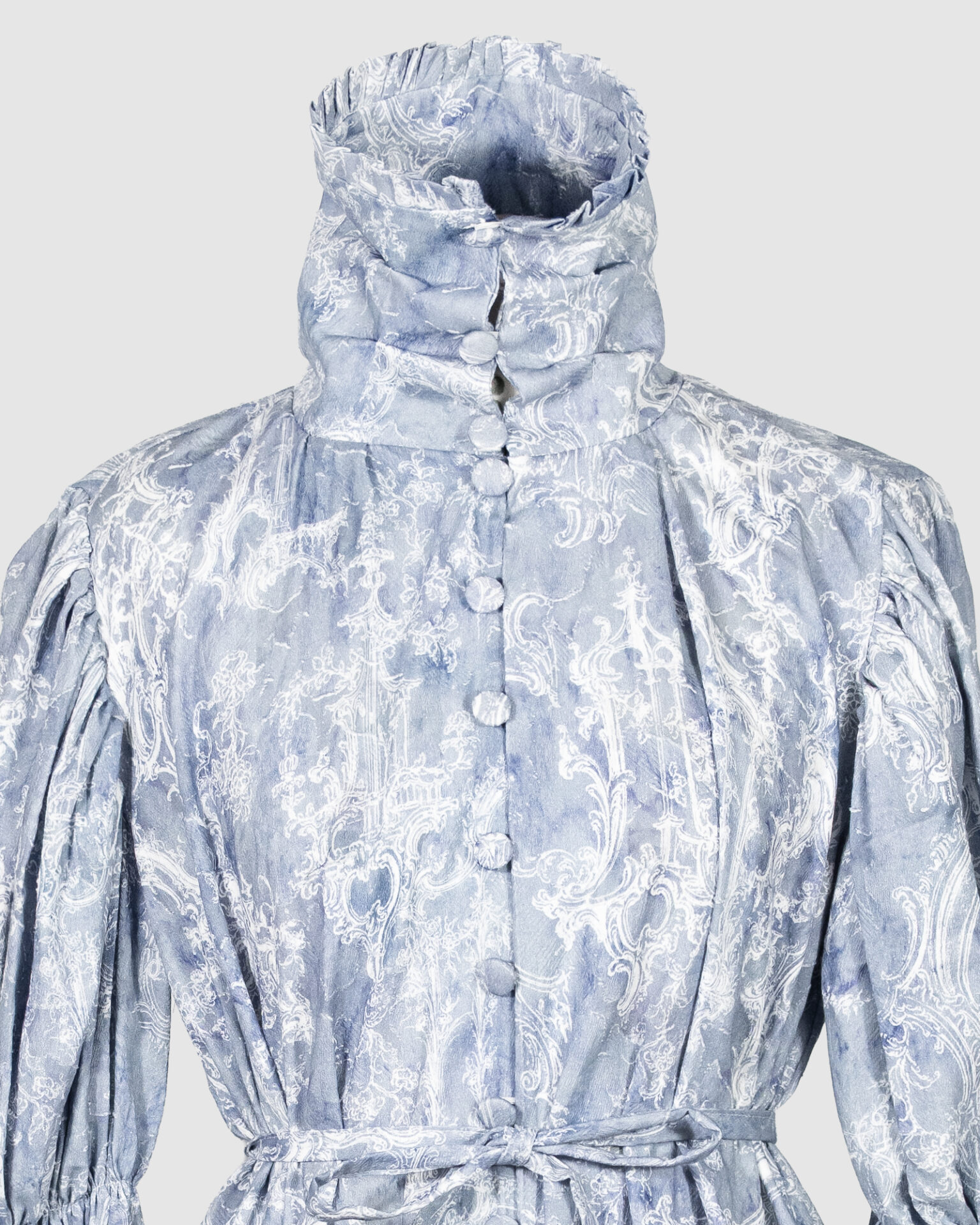 This loose-fitting blouse features a softly pointed front hem, a stand collar accented with a short ruffle, and a front button closure.
